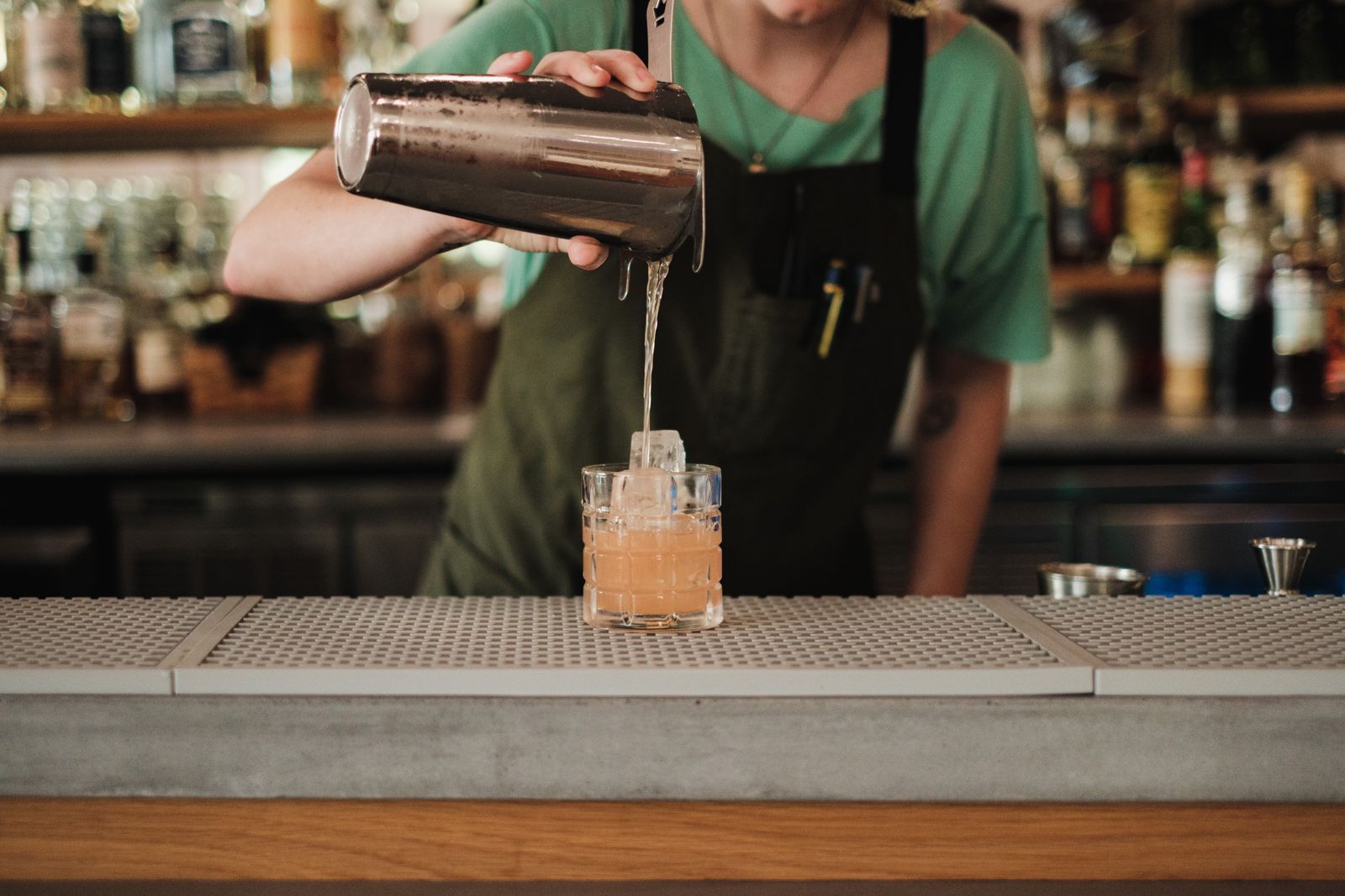 A bartender in a green t-shirt and dark apron pouring a shaken cocktail from a metal shaker into a glass over ice, with shelves of bottles blurred behind the bar counter