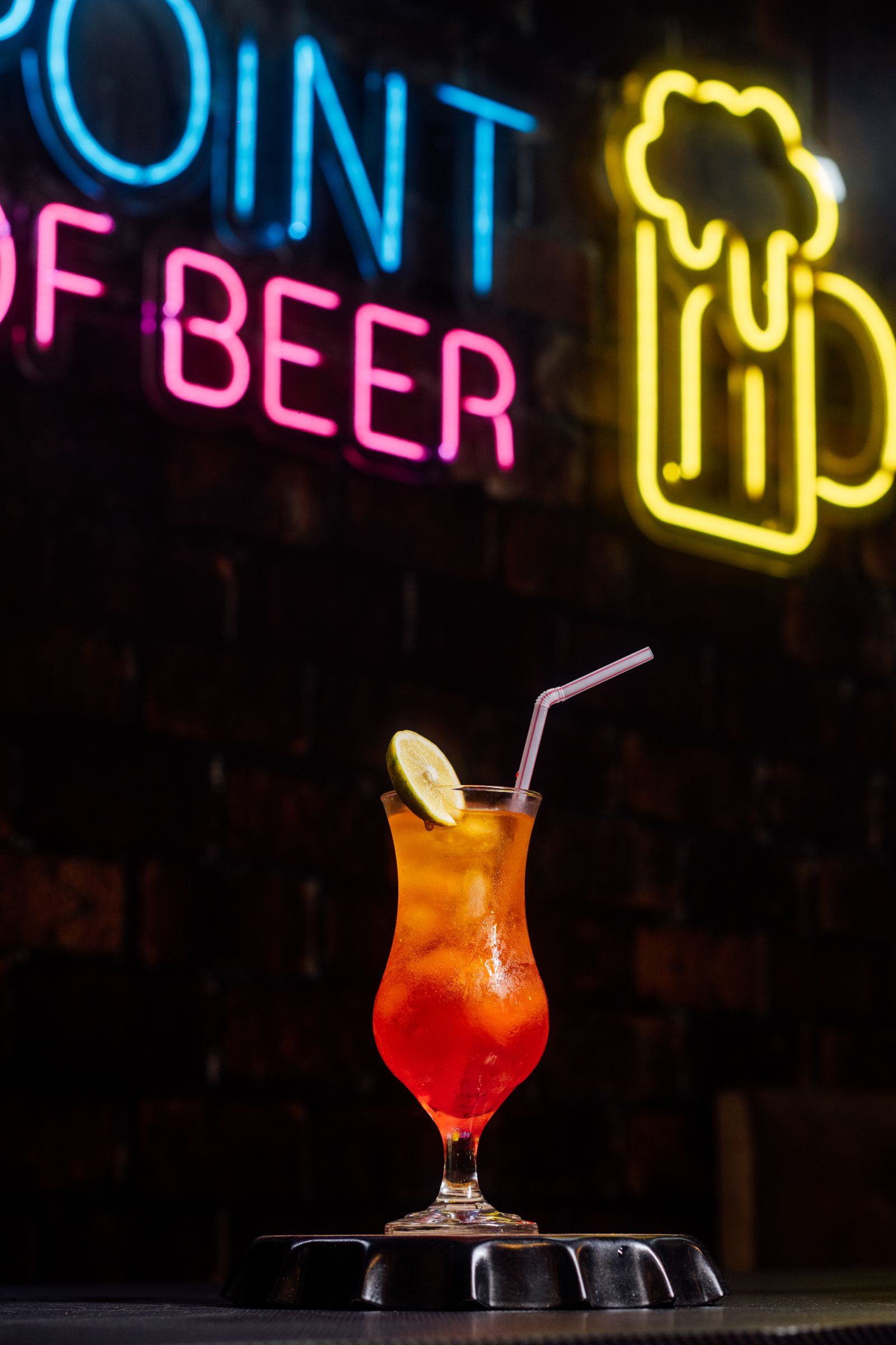 Close-up of an orange cocktail with lime garnish on a bar counter with out-of-focus pink and blue neon signage in the background