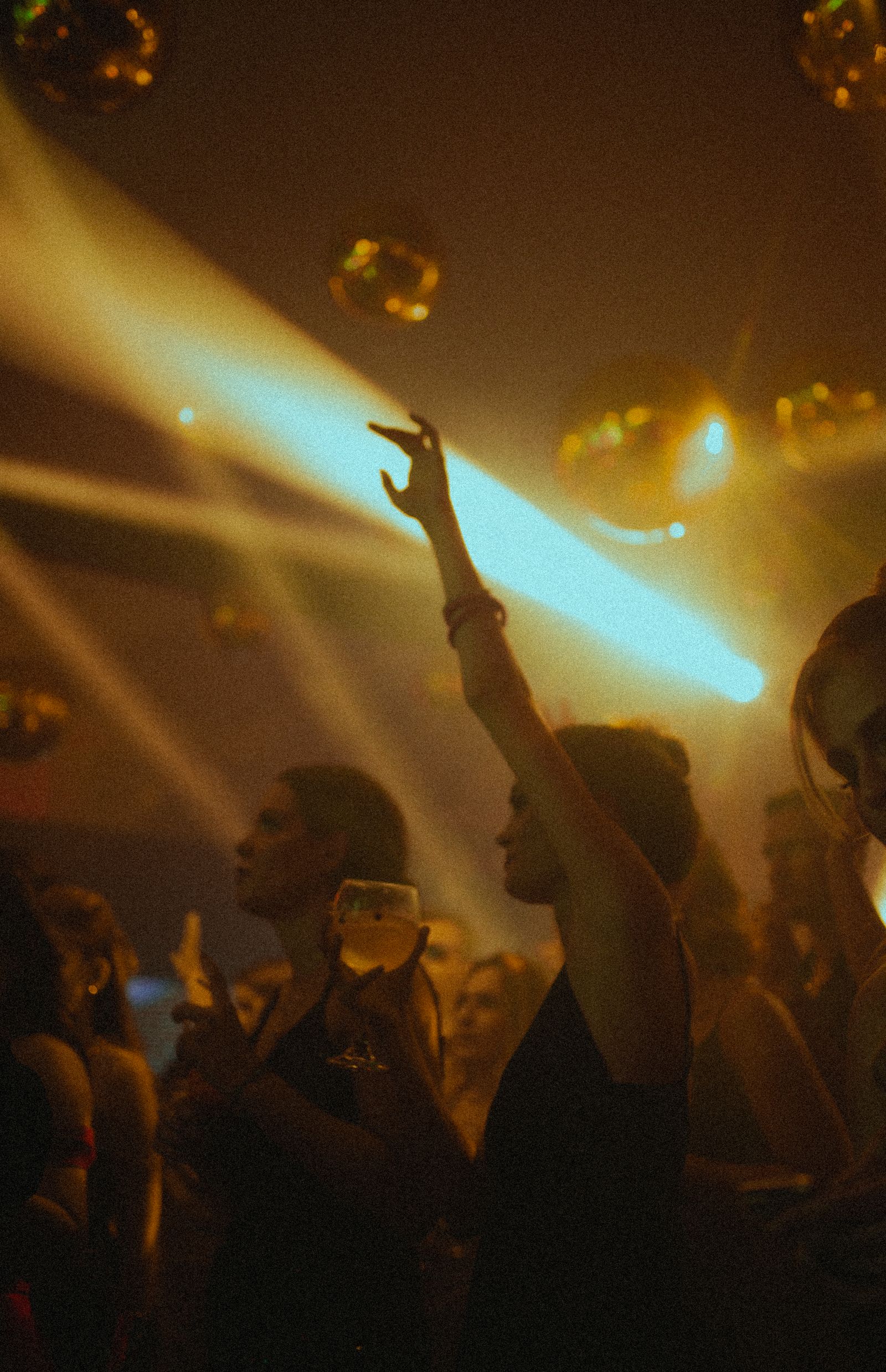 Warm gold-toned hazy shot of a small silhouetted crowd, one person with arm raised holding a drink glass, with a cyan light beam crossing the frame