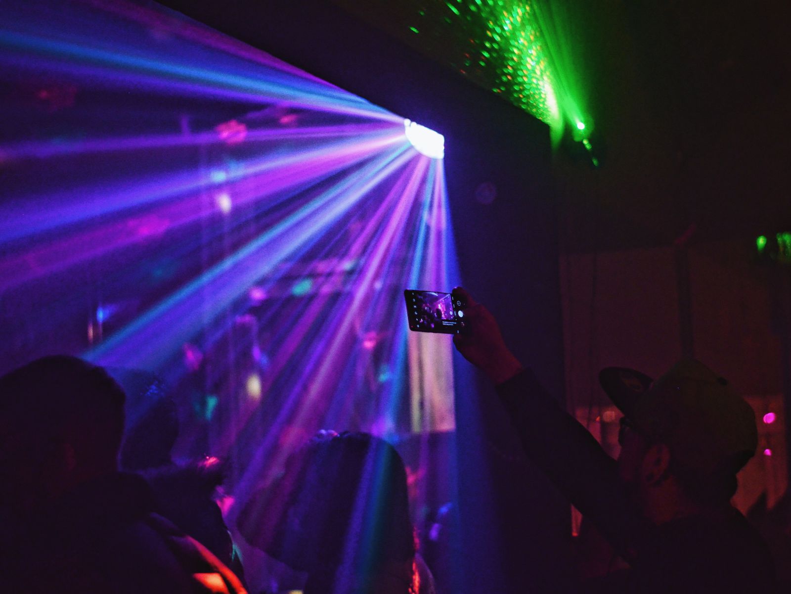 Purple and green laser beams fanning across a dark room with a silhouetted crowd in the foreground and one person holding up a phone to film