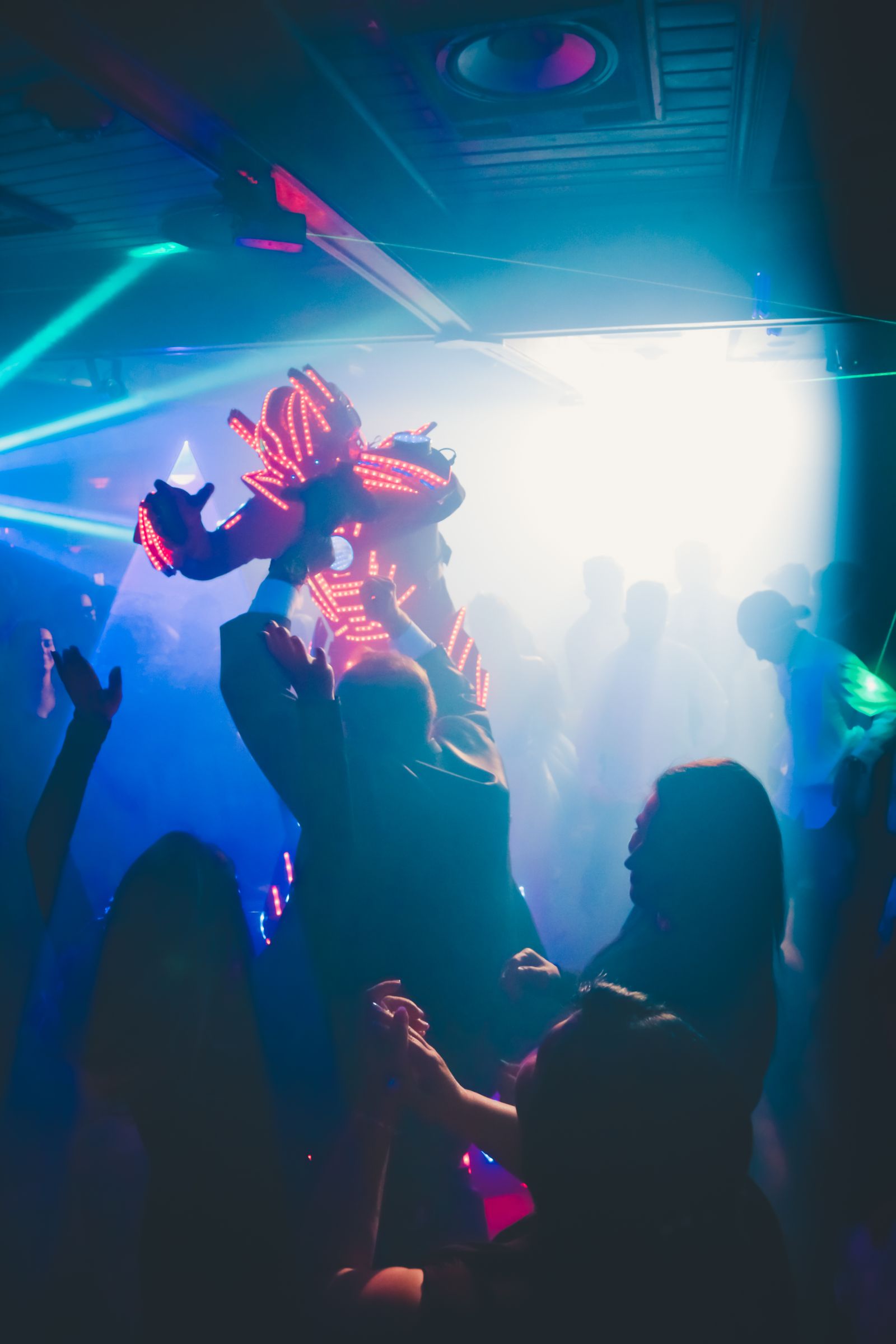 Silhouetted crowd with raised arms dancing under blue and teal laser beams in a hazy club atmosphere